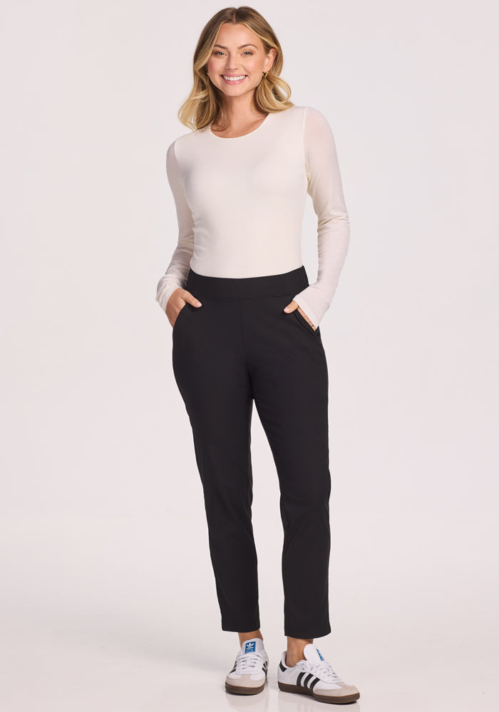 A woman smiles with hands in her pockets, wearing a long-sleeve white top, Woolx Nicole Petite Pants in black, and white sneakers with brown soles against a plain light background. 