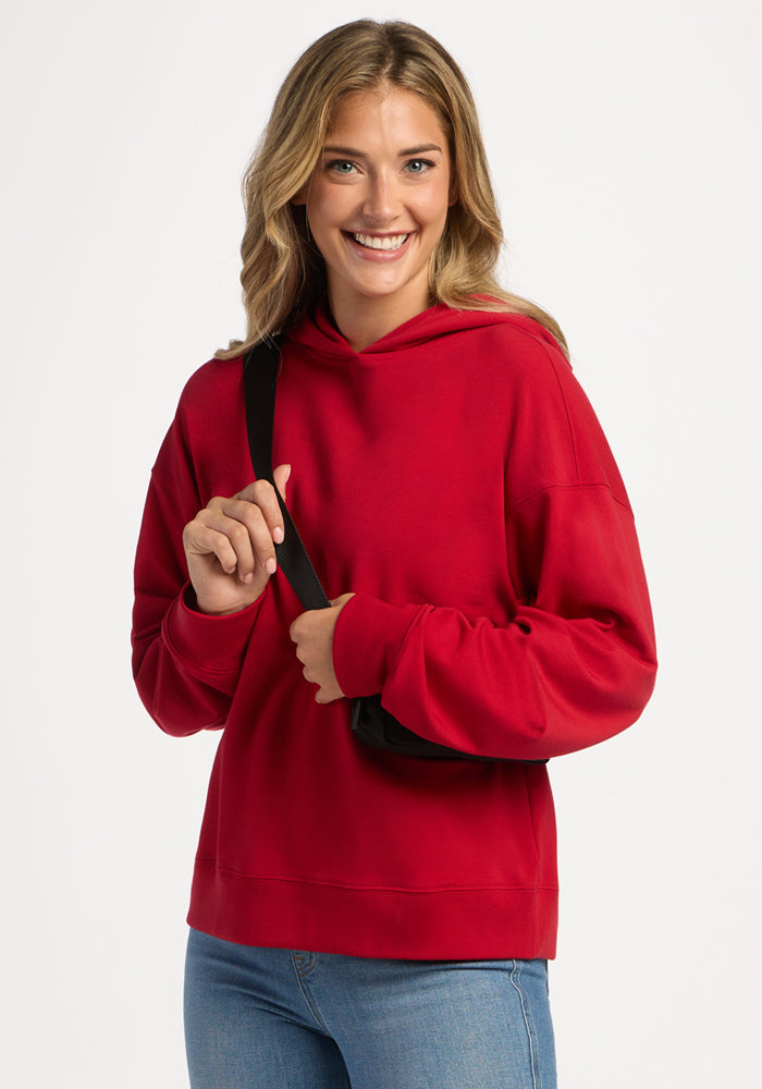 Smiling woman wearing a bright red Woolx Genevieve Hoodie - Maraschino Cherry and a black crossbody bag.