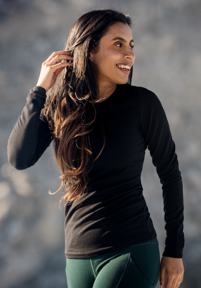 A woman wearing the Woolx Sadie Baselayer Top in black and dark green pants, crafted from Duralite merino fabric, smiles as she looks to her right while standing outdoors against a blurred gray background.