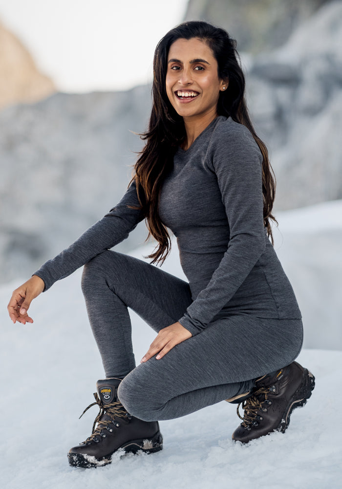 A woman wearing Woolx Stella Leggings in Cinder Melange and a gray long-sleeve top kneels on snowy ground, smiling at the camera, with a blurred outdoor winter background.