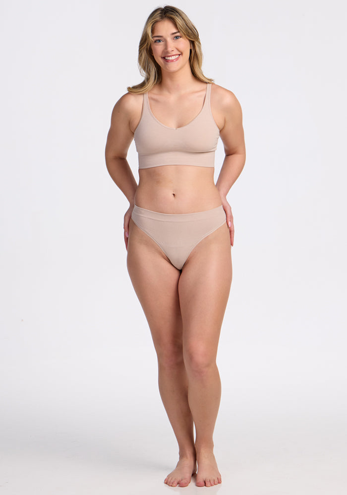 A woman with long blonde hair stands barefoot on a white background, smiling. She is wearing the Woolx Farrah Thong in Sand, part of a matching set of seamless beige underwear.