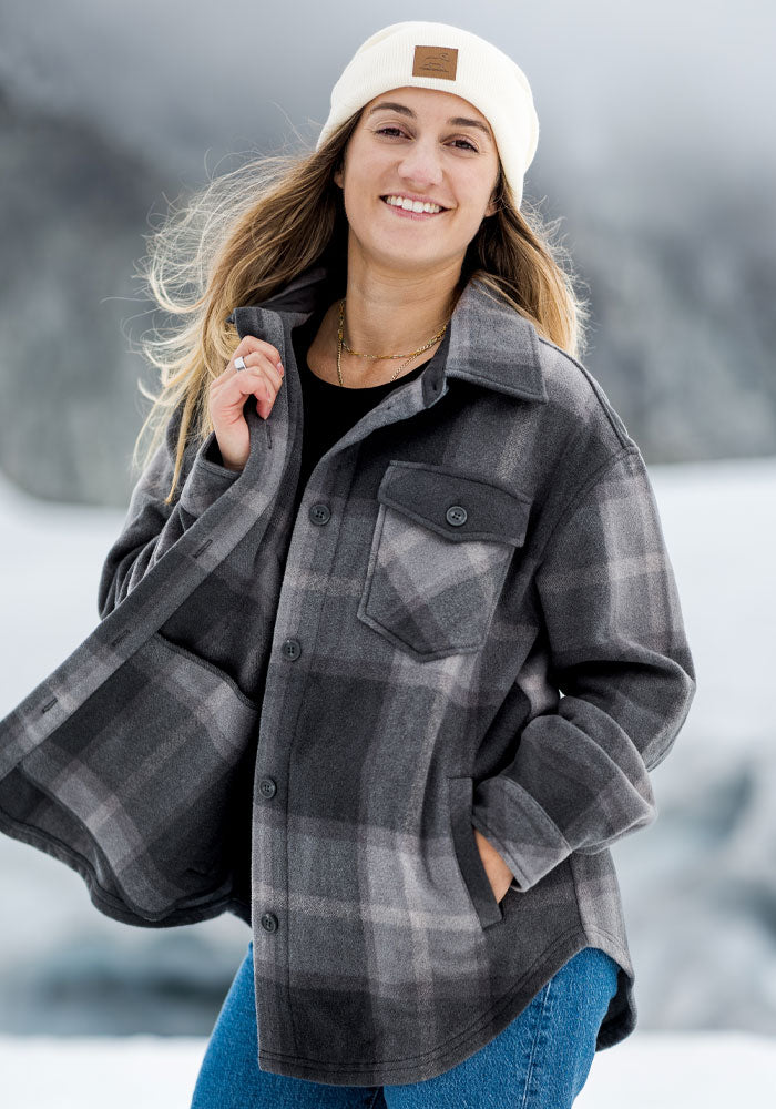 Smiling woman in Woolx Sawyer Shacket - Volcanic Ash Plaid and white beanie stands outdoors in snowy mountains.