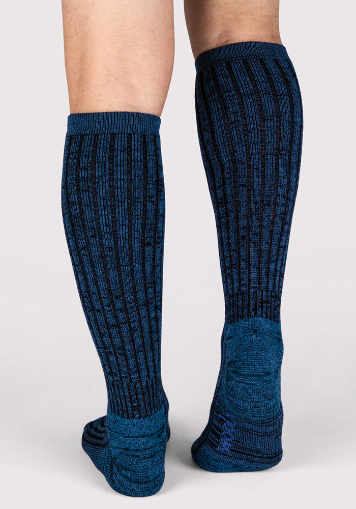 A person stands on a light grey background, showing the back view of their legs in cozy Woolx Mens Joey Mid Calf Socks in steel blue, highlighting the detailed blue and black pattern of these Merino wool socks.