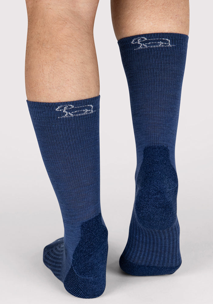A close-up of the back of a person's lower legs wearing Woolx Mens Harbor Crew Sock in Sailor Blue, featuring moisture-wicking fabric, a ribbed texture, and a small white logo near the top against a plain light background.