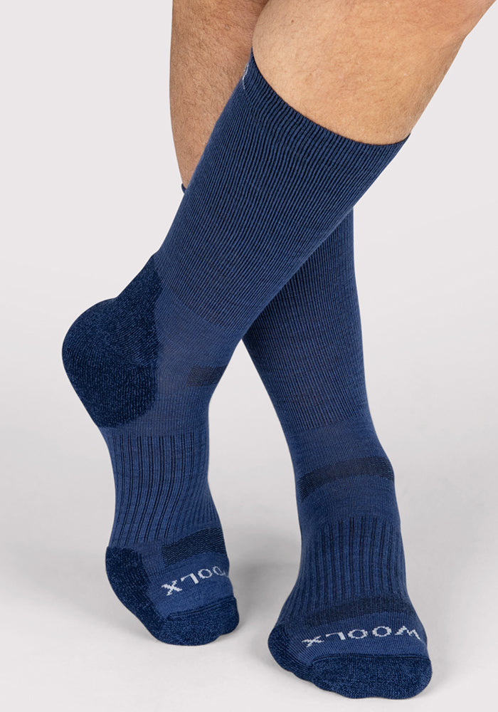A person wearing Woolx Mens Harbor Crew Sock in Sailor Blue, featuring ribbed texture and reinforced heels and toes, stands with legs crossed against a light gray background.