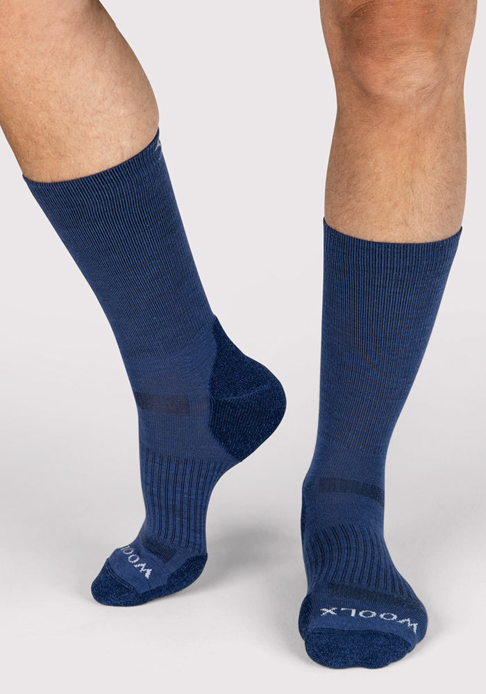 A person wearing Woolx Mens Harbor Crew Socks in Sailor Blue stands on a light surface. The tall Merino wool socks have ribbed texture, cushioned heels and toes, with "WOOLX" visible on the soles.