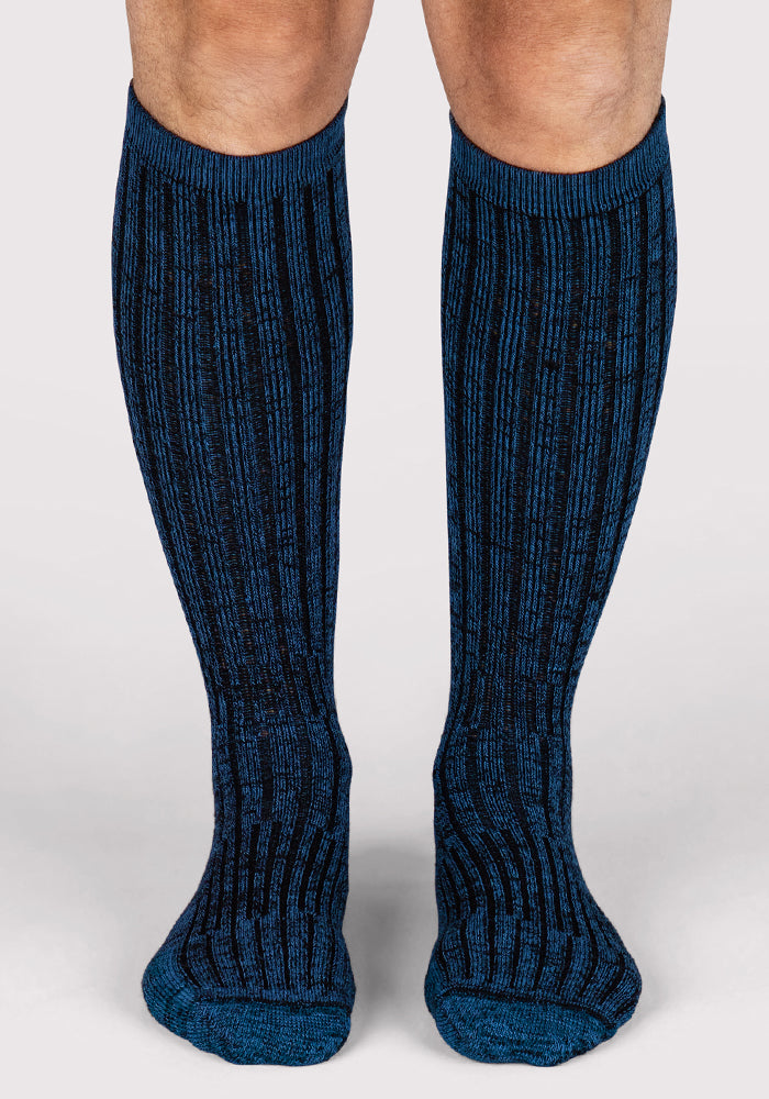 A person wears Woolx Mens Joey Mid Calf Socks in Steel Blue, standing on a light, neutral background with only their lower legs and feet visible.