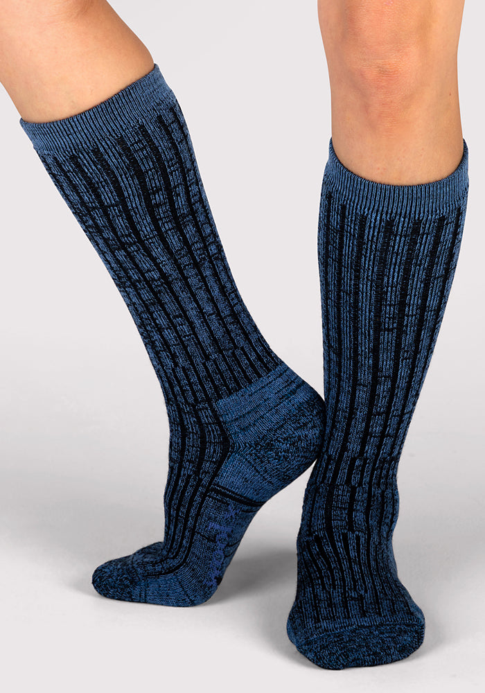 A person stands on a light gray surface wearing Woolx Womens Joey Mid Calf Socks in Steel Blue, one foot flat and the other slightly raised to display both the side and front of the socks.