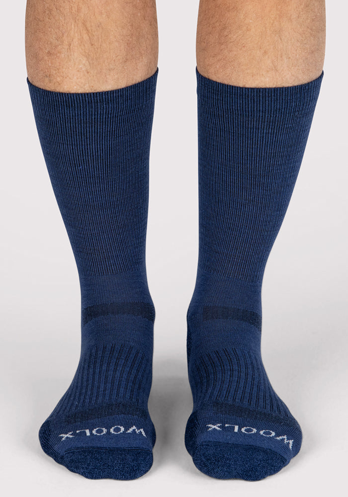 A pair of feet wearing Woolx Mens Harbor Crew Socks in Sailor Blue, mid-calf Merino wool with "WOOLX" in white on the toes, stands on a light surface. These moisture-wicking socks offer comfort and style.