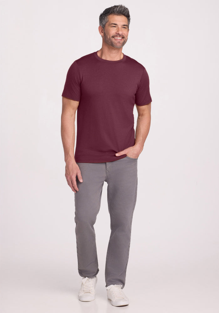 A smiling man with short dark hair and a beard wears the Woolx Endurance Tee in Grape Harvest, paired with gray pants and white sneakers, standing against a plain light background.