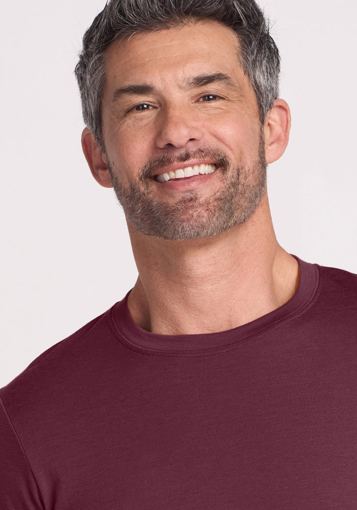 A middle-aged man with short gray hair and a salt-and-pepper beard smiles while wearing the Woolx Endurance Tee in Grape Harvest against a plain light background.