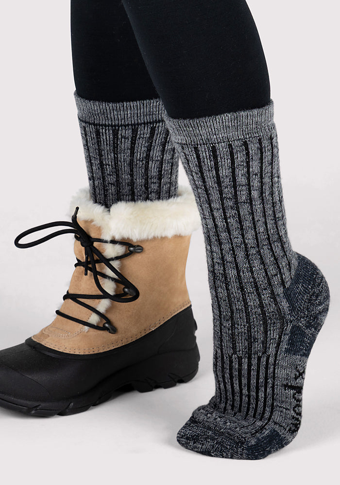 A person wearing Woolx Womens Joey Crew Socks in Stone Grey and tan winter boots with black laces and white fur trim, standing against a plain light background.