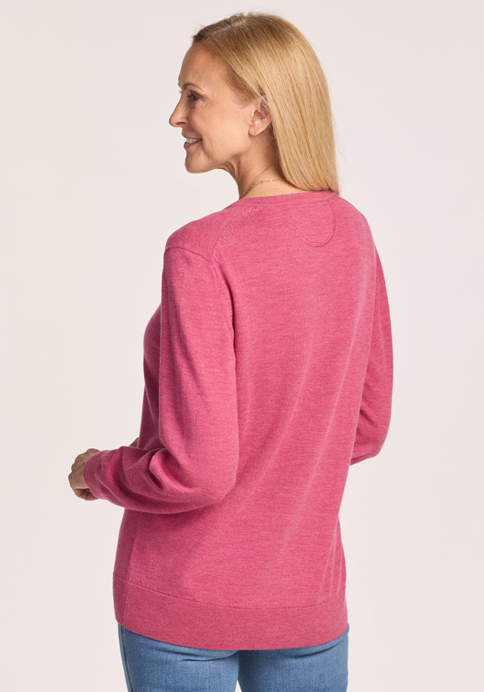 A woman with straight blonde hair wears the Woolx Francesca Sweater in Wild Salmon and blue jeans, standing slightly turned with a smile against a plain light background.