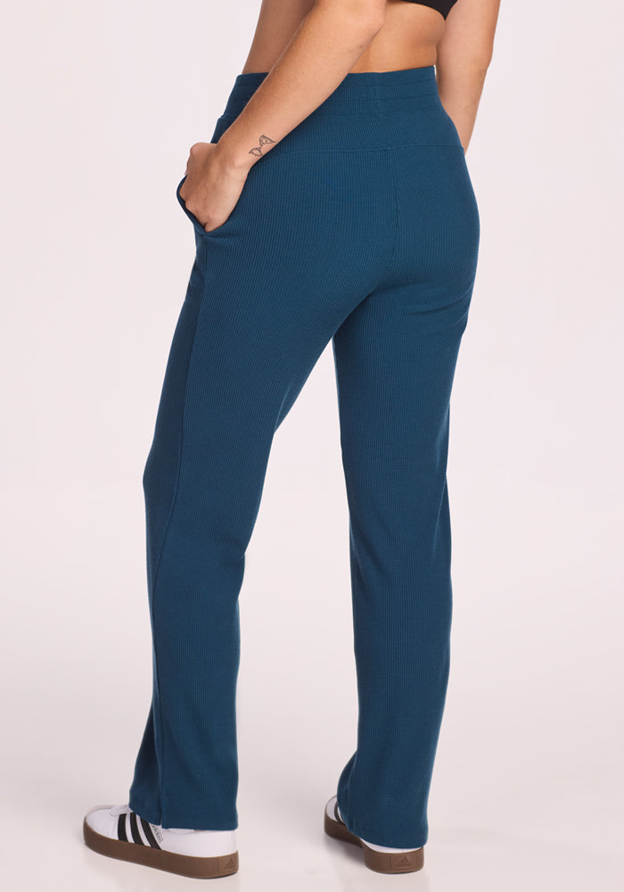 A person wearing Woolx Luca Ribbed Pants in Majolica Blue and a black crop top stands with one hand in their pocket, facing away. They're wearing black and white sneakers, with a small tattoo visible on their forearm.