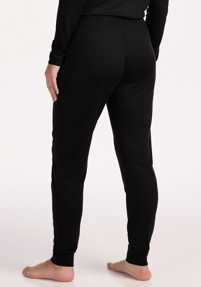 Person shown from the waist down, back view, standing barefoot on a light surface in Woolx Mary Kate Lounge Pants - Black with a matching long-sleeve top—ideal travel loungewear offering moisture-wicking comfort.