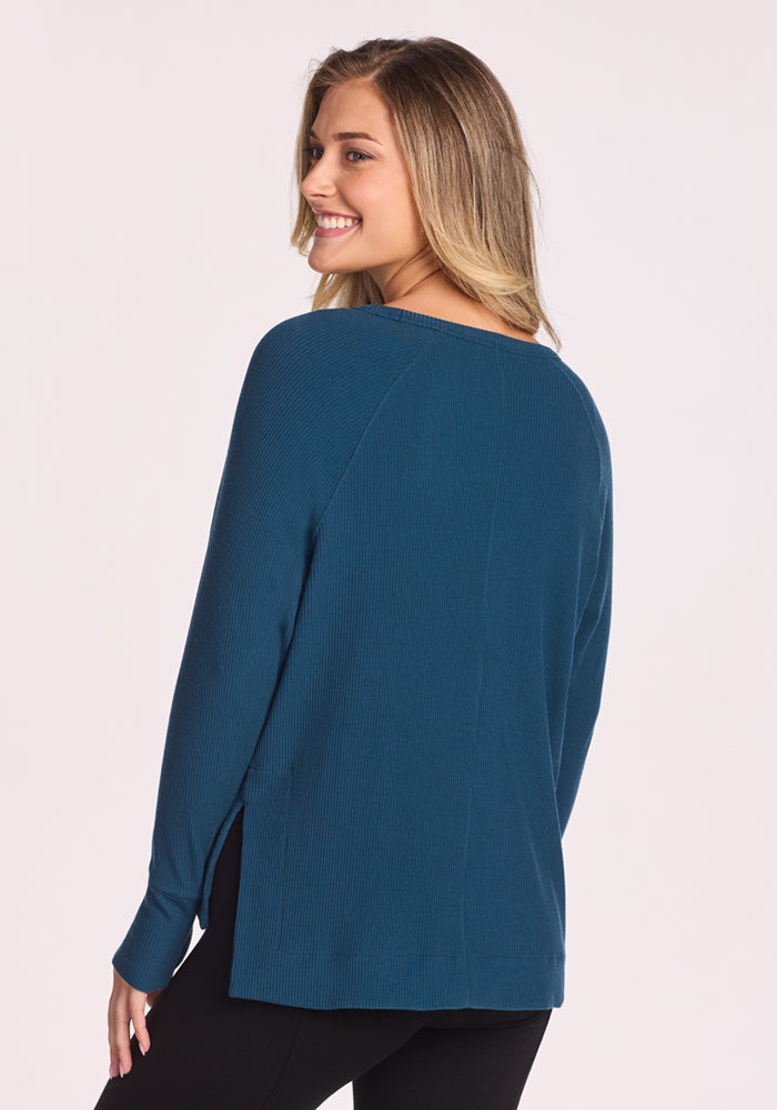 A woman with long blonde hair smiles over her shoulder, wearing the Woolx Ainsley Sweater in Majolica Blue—crafted from temperature-regulating Merino wool—paired with black pants against a plain light background.