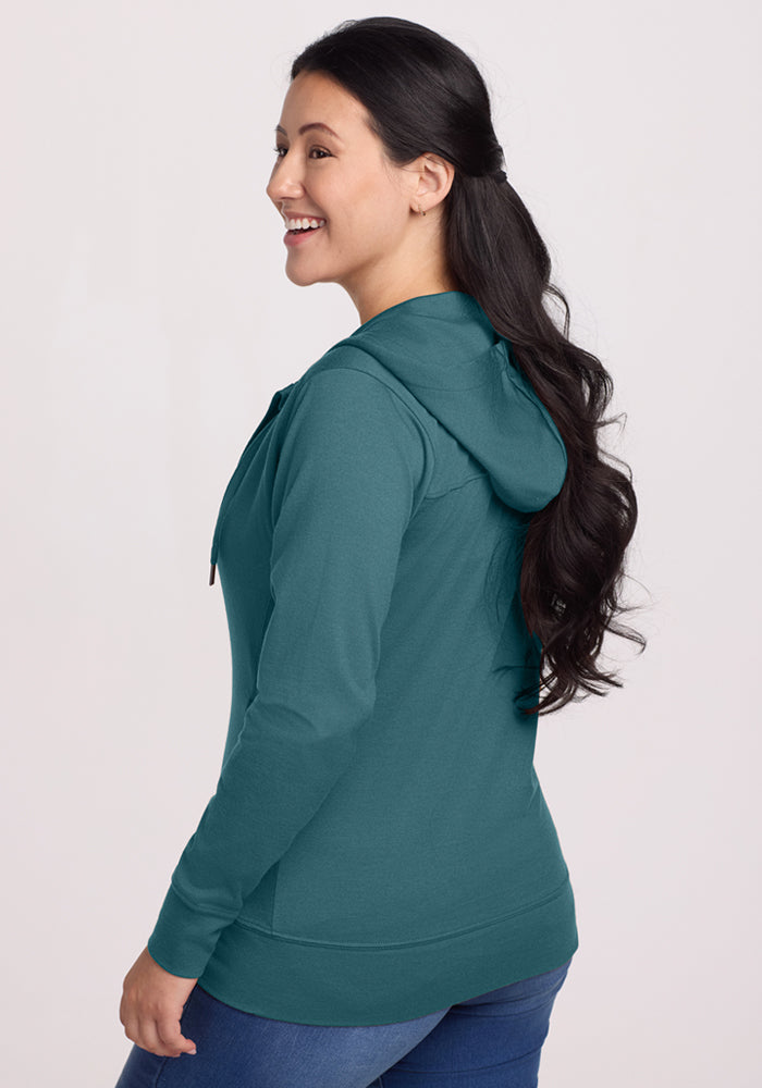 A woman with long dark hair in loose waves smiles while wearing the Woolx Ryann Hoodie - Tidepool and blue jeans, turned to the side against a plain light background—an ideal travel essential for comfort and style.