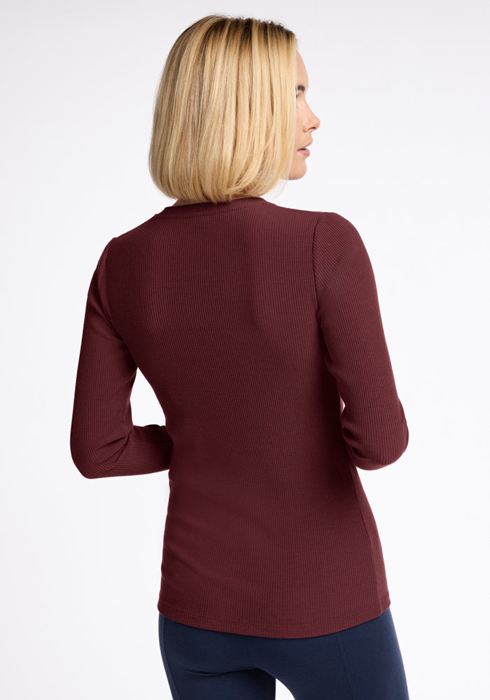 A woman with straight blonde hair, seen from the back, wears the Woolx Hadley Ribbed Crew in Toasted Garnet and dark pants—an ideal lightweight layering piece for travel—set against a plain white background.
