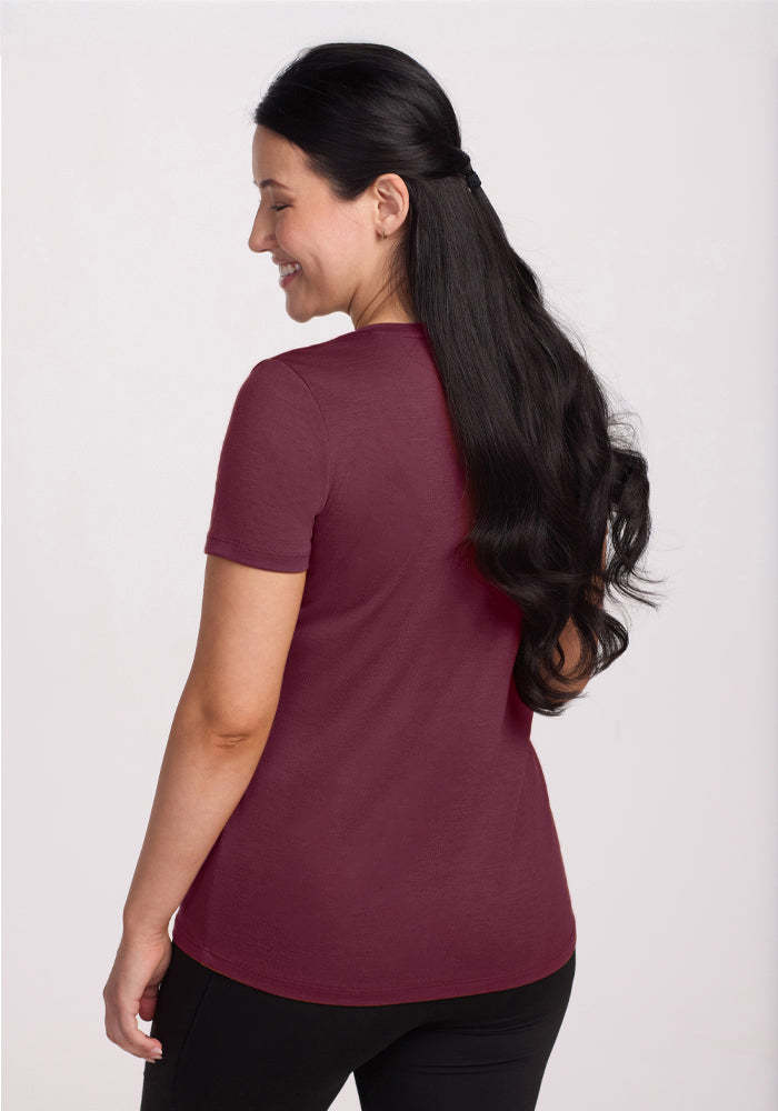 A woman with long, dark wavy hair stands facing away, wearing the Woolx Junie V Neck in Grape Harvest and black pants, smiling slightly against a plain light background.