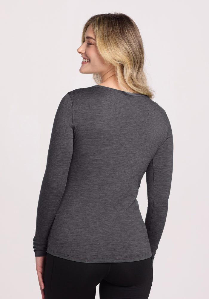 A blonde woman smiles over her shoulder, wearing the Woolx Remi Long Sleeve T-Shirt in Cinder Melange and black pants. The odor-resistant top stands out against the plain, light background.