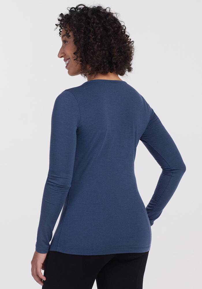 A woman with short curly hair wears the Woolx Remi Long Sleeve T Shirt – Starry Night and black pants, standing smiling, shown from the back and slightly turned to the side against a plain light background.