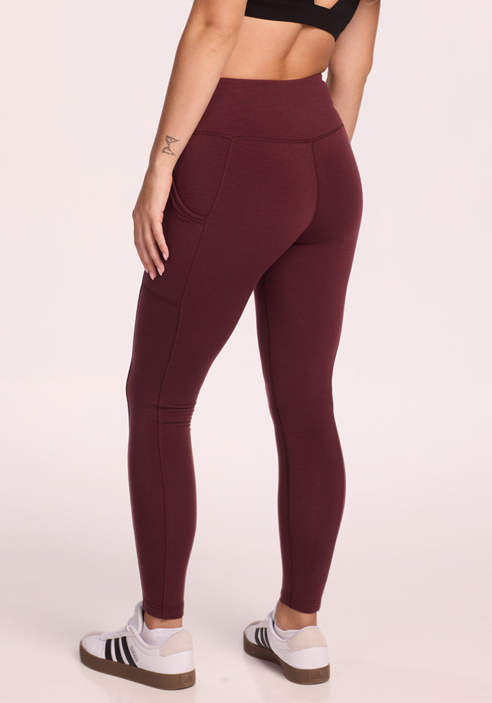 A person is shown from the back wearing Woolx Piper Pocket Leggings in Toasted Garnet, a black sports bra, and white sneakers with black stripes, standing against a neutral background with their left arm relaxed by their side.