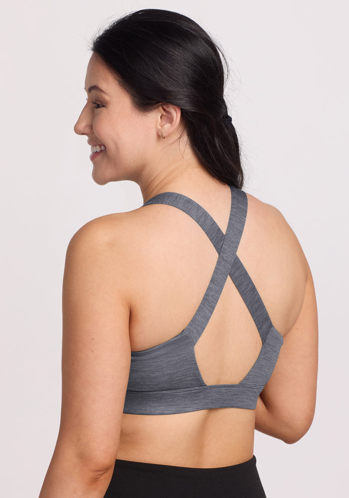 A woman with long dark hair in a ponytail smiles while wearing the Woolx Livi Bra in Graphite Heather, featuring crisscross back straps. She is shown from behind against a plain light background.