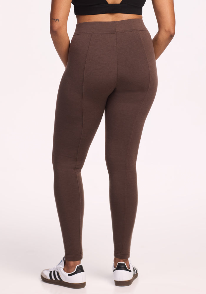 A person stands facing away, wearing Woolx's Stella Petite - Bracken high-waisted leggings in brown, a black crop top, and white sneakers with black stripes against a plain light background.