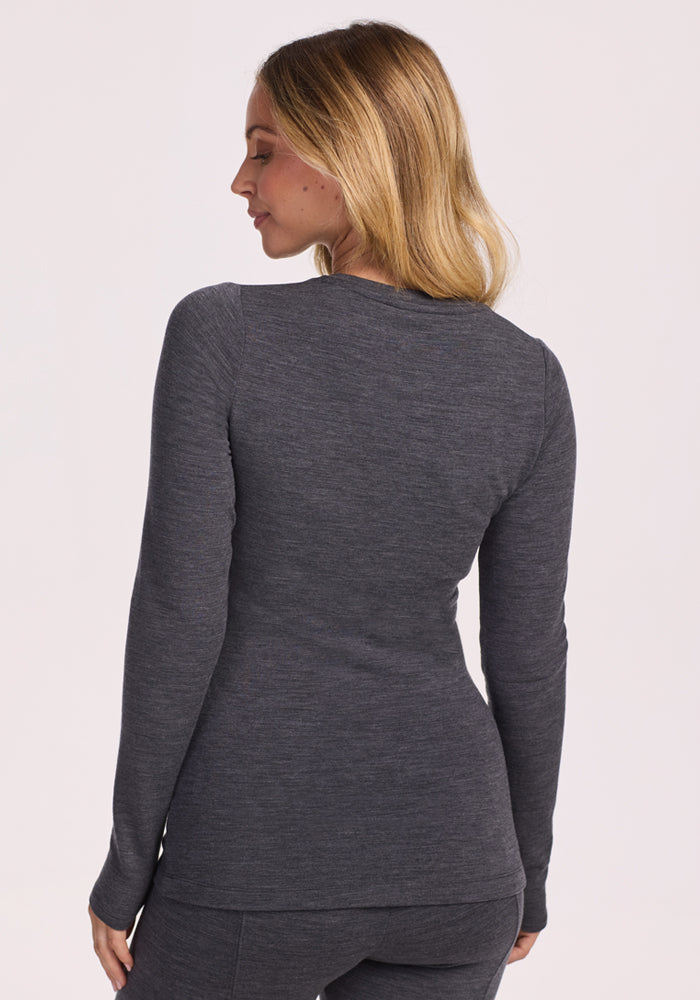 A blonde woman, wearing the Woolx Sadie Baselayer Top in Cinder Melange, stands facing away from the camera with her head turned slightly to the side.