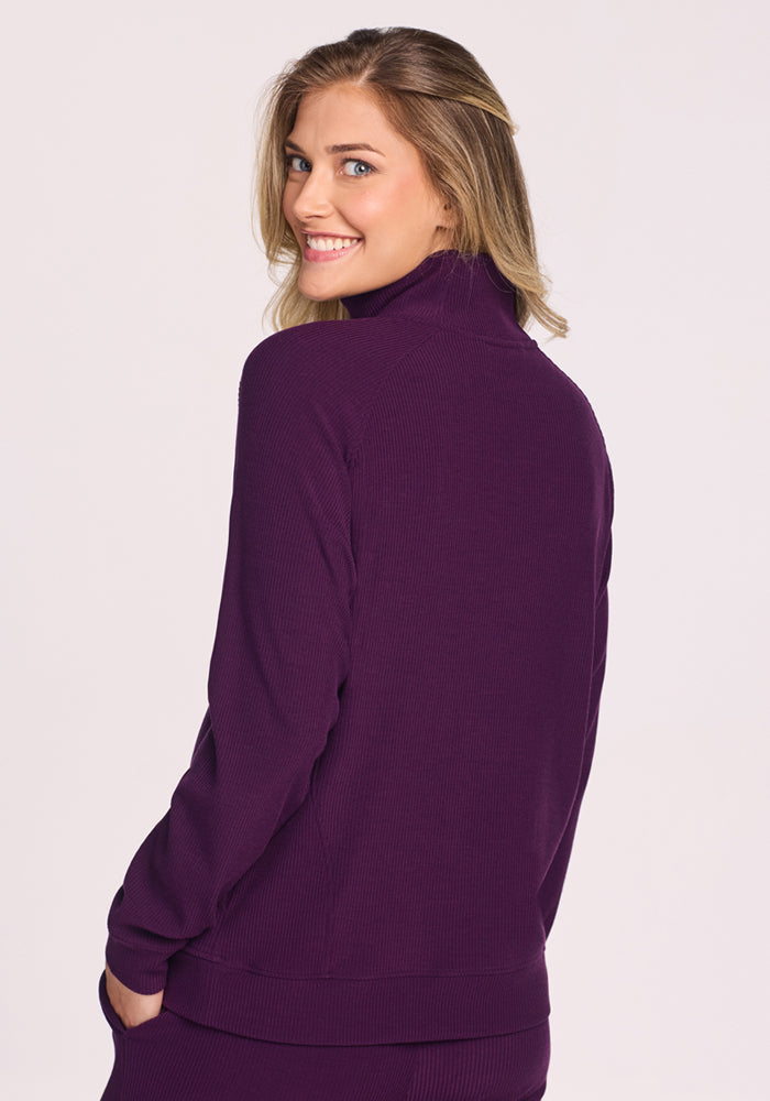 A woman with blonde hair smiles over her shoulder while wearing the Woolx Quinn Ribbed 1/4 Zip in Majestic Purple. She faces away against a plain light gray background.