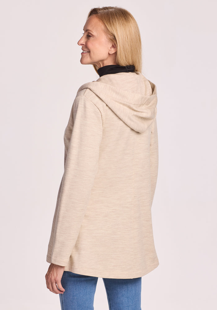 A woman with blonde hair in blue jeans is standing and smiling, shown from the back and side, wearing the Woolx Edna Hooded Cardigan in Cream Heather against a plain light background.