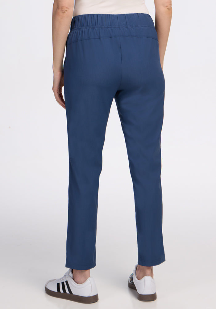 A person stands facing away from the camera, wearing Woolx Nicole Ankle Pants in Vintage Indigo with an elastic waistband and white sneakers with black side stripes, set against a plain white background.