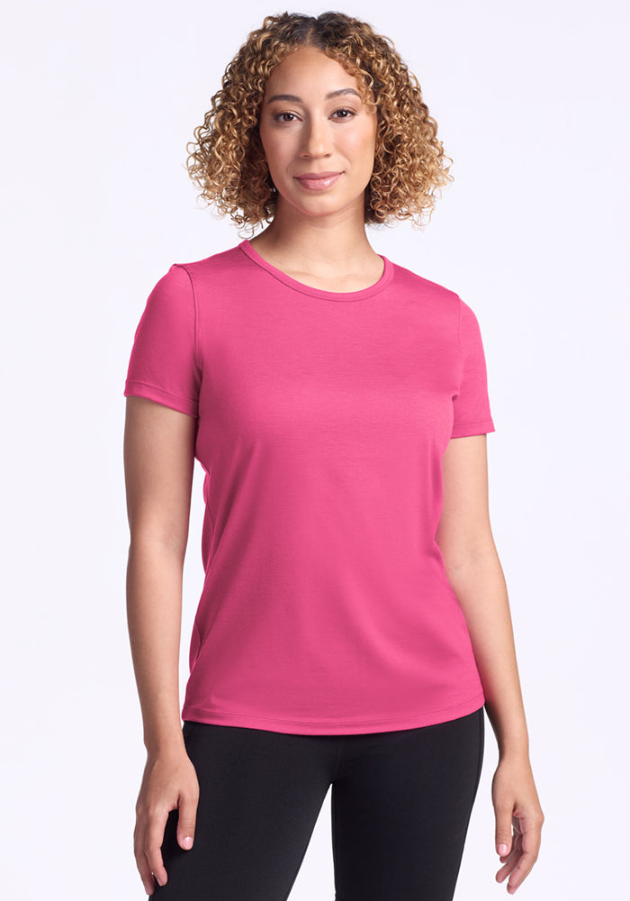 A person with curly hair stands facing forward against a white background, wearing the Woolx Liza Crew Neck Tee in Raspberry Fizz and black pants. 