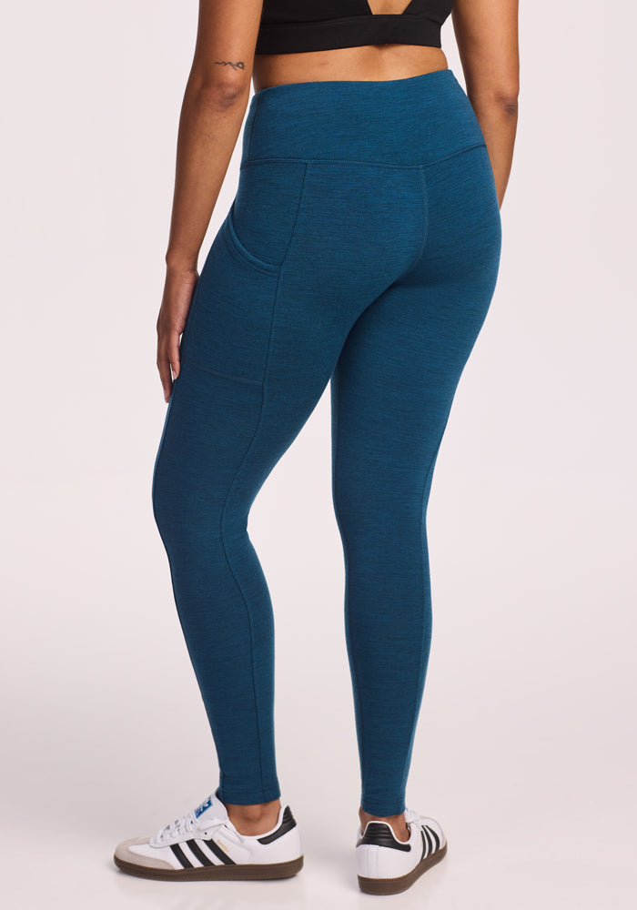 A person stands with their back to the camera against a plain light background, wearing a black crop top, Woolx Piper Pocket Leggings in Majolica Blue, and white sneakers with black stripes.