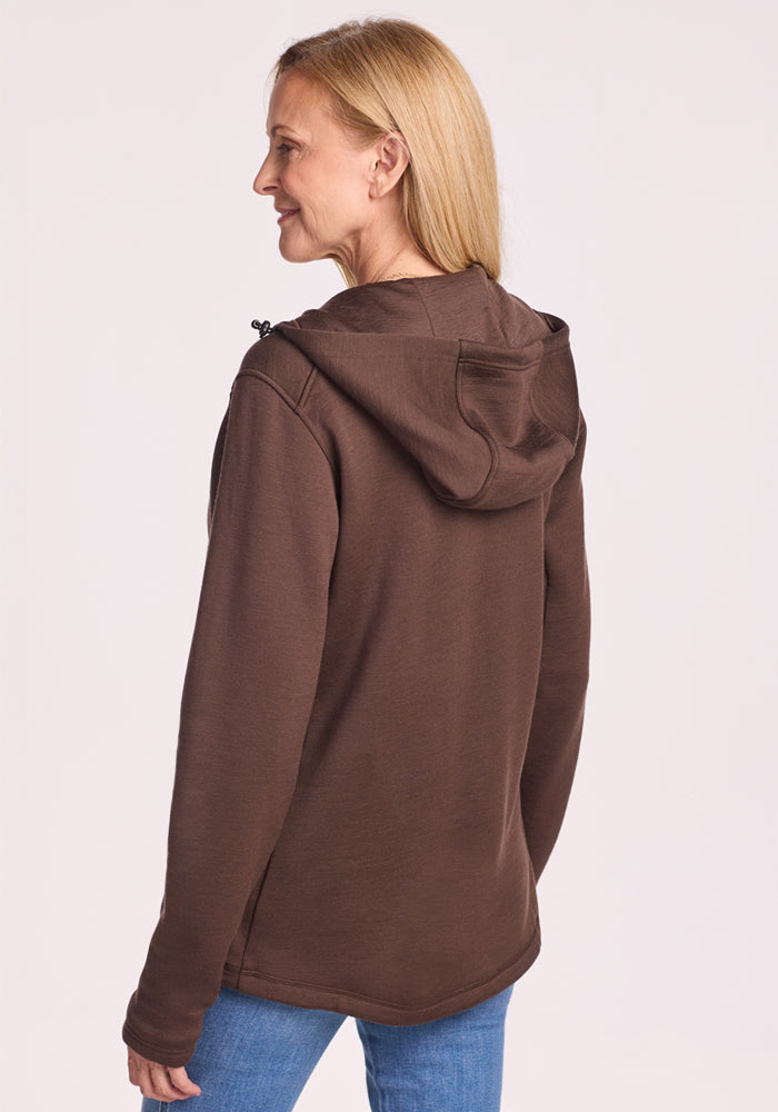 A woman with blonde hair wears the Woolx Cubby Hooded Sweatshirt in Bracken and blue jeans, standing against a light background, smiling as she faces away with her head turned slightly to the side.