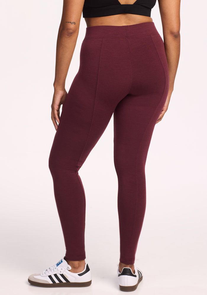 A person wearing a black crop top, Woolx Stella Leggings in Toasted Garnet, and white Adidas sneakers with black stripes stands with their back to the camera against a plain light background.