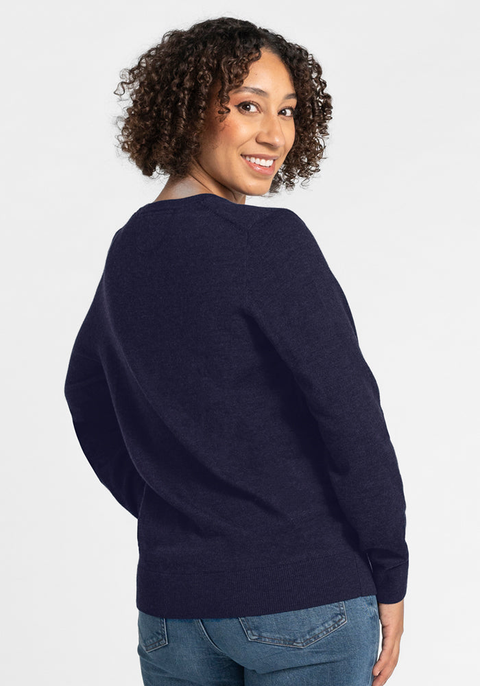 A woman with curly hair wears the Woolx Francesca Sweater in Storm Cloud and blue jeans, standing against a white background and looking back over her shoulder with a smile.