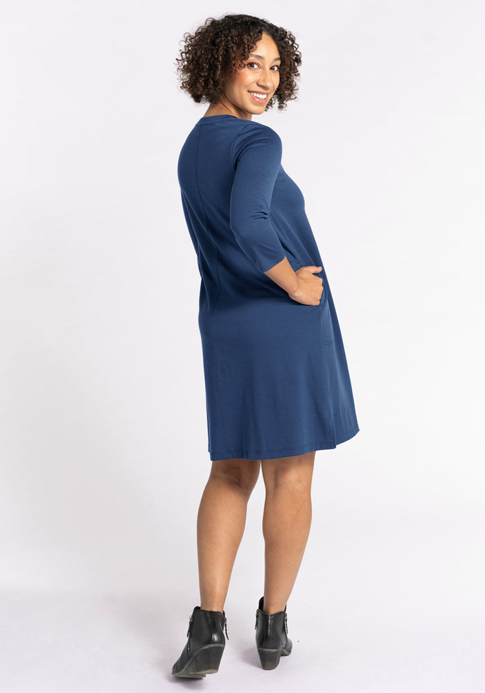 A woman with curly hair smiles over her shoulder, wearing the Woolx Rissa A Line Dress - Starry Night. This knee-length merino wool dress with three-quarter sleeves and pockets is perfect for layering in fall and winter. She stands in ankle boots on a white background.