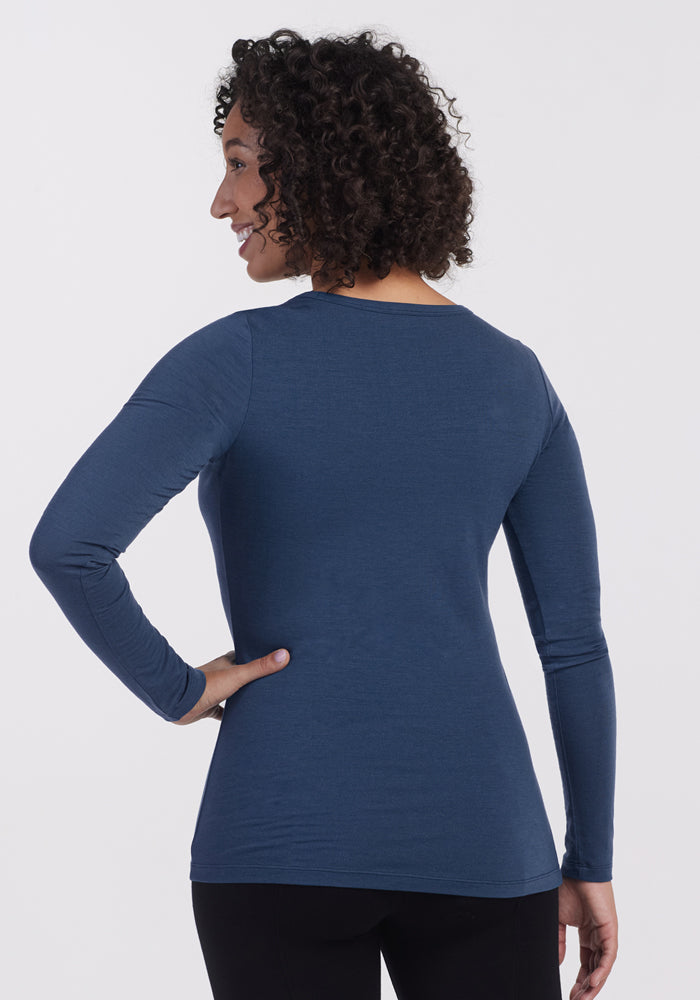 A woman with curly hair faces away from the camera, wearing the Woolx Layla V Neck Top in Starry Night with black pants, standing against a plain white background.