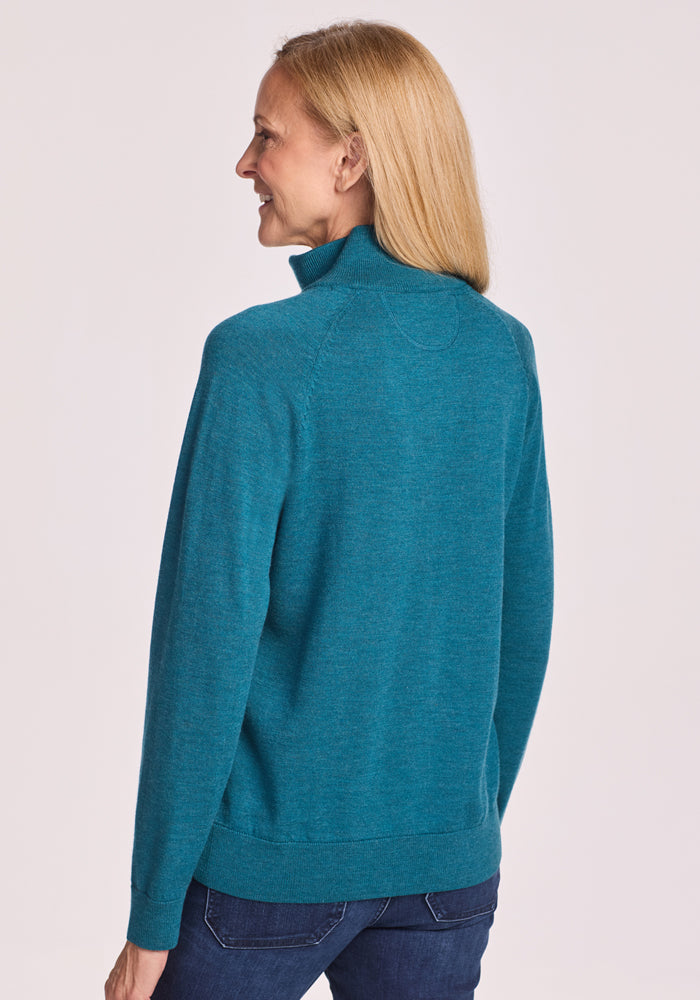 A woman with straight blonde hair is shown from the back, smiling as she looks right. She wears the Woolx Collins Half Zip Sweater in Harbor Teal and blue jeans, set against a light background.