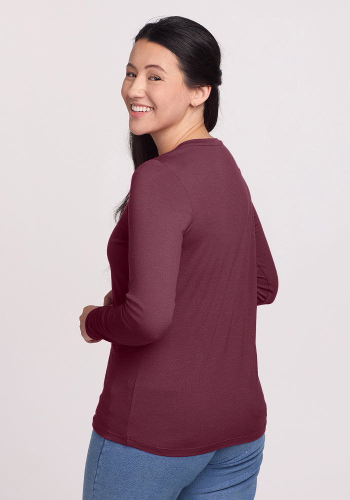 A woman with long dark hair, wearing the Woolx Millie Long Sleeve in Grape Harvest and blue jeans, smiles as she turns to look over her shoulder against a plain light background.