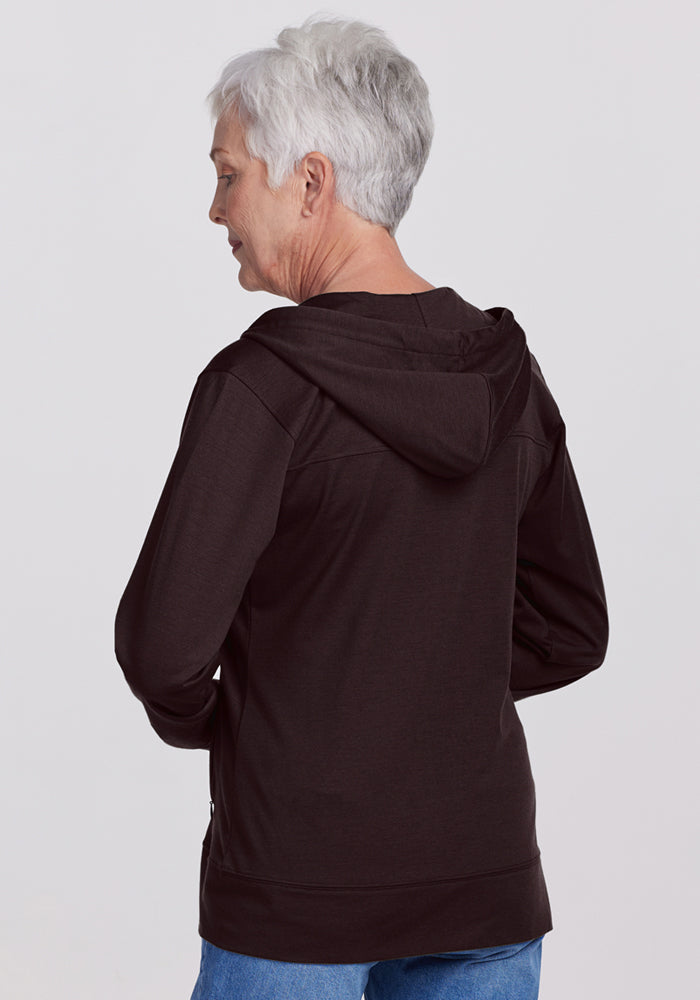 An older woman with short gray hair is seen from behind, wearing the Woolx Ryann Hoodie in French Roast and blue jeans, standing against a plain light background.