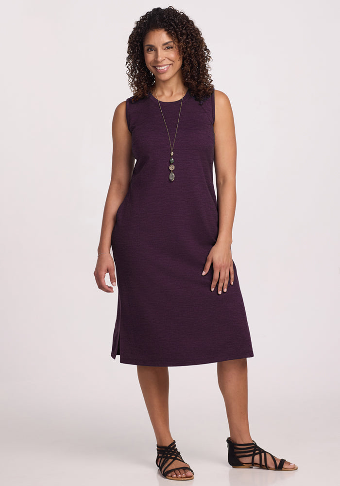 A woman with curly hair smiles while wearing the Woolx Cassie Dress in Deep Plum, paired with black strappy sandals and a long necklace, standing against a plain white background. 