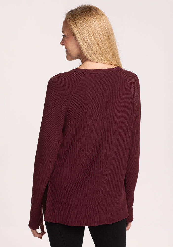 A woman with long blonde hair, seen from behind, wears black pants and a Woolx Ainsley Sweater in Toasted Garnet against a plain white background.