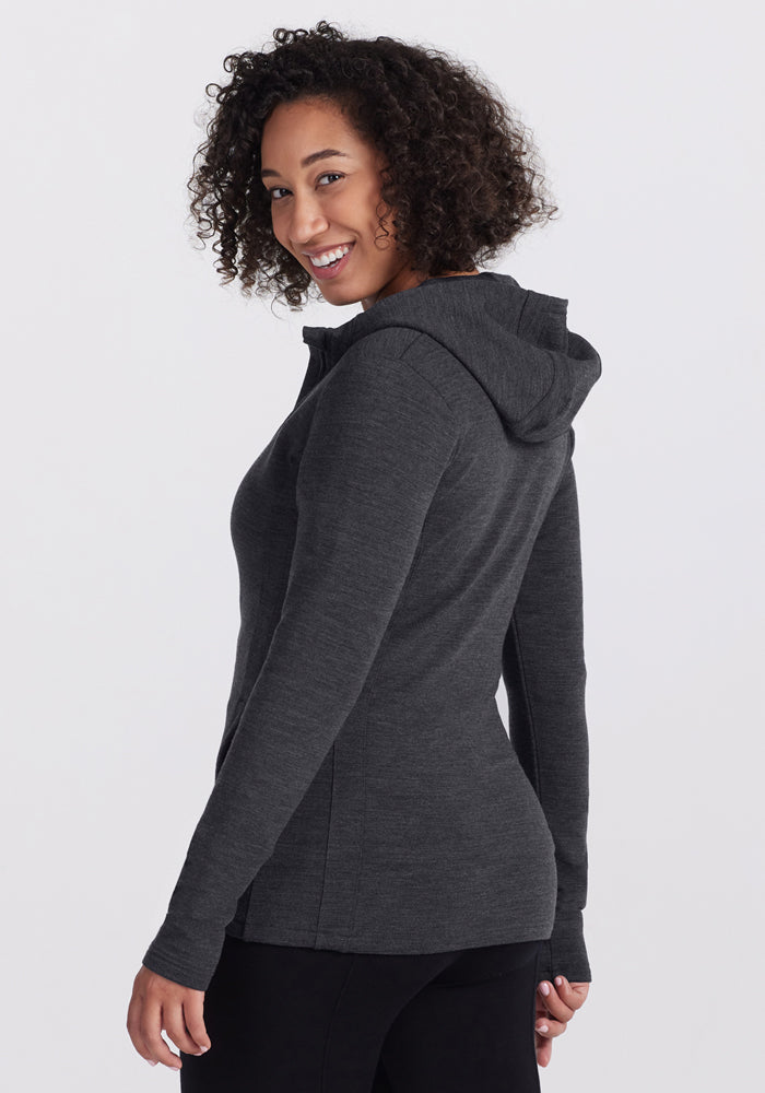 A person with curly hair, wearing a Woolx Zoey Zip Up Hoodie in Cinder Melange, stands turned slightly to the side and smiles over their shoulder against a plain light background.