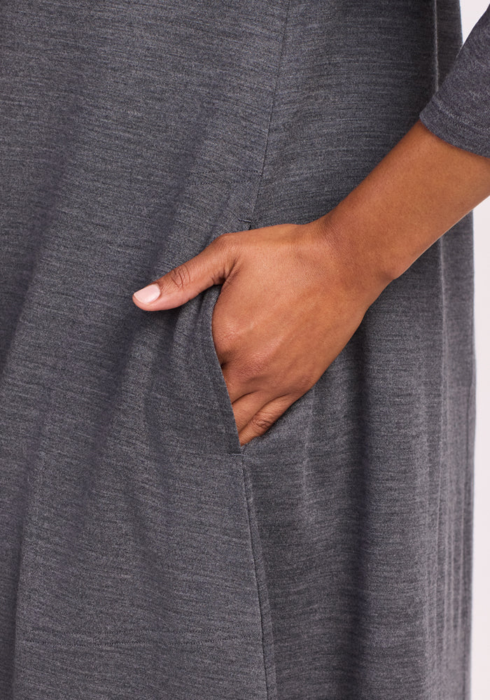 A close-up shows a hand in the side pocket of the Woolx Rissa A Line Dress in Cinder Melange, highlighting its soft Merino wool texture and cozy sleeve—perfect for fall and winter—against a plain light backdrop.