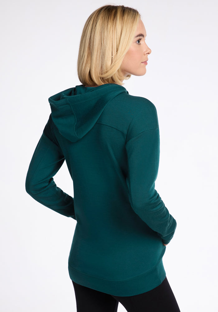 A woman with blonde hair, wearing the Woolx Callie Hoodie in Riverbank Green and black pants, stands with her back to the camera and looks to the side against a plain white background.