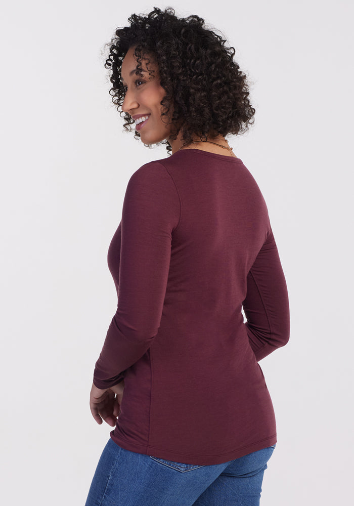 A woman with curly hair, wearing the Woolx Remi Long Sleeve T Shirt in Grape Harvest and blue jeans, stands sideways and smiles against a plain white background.