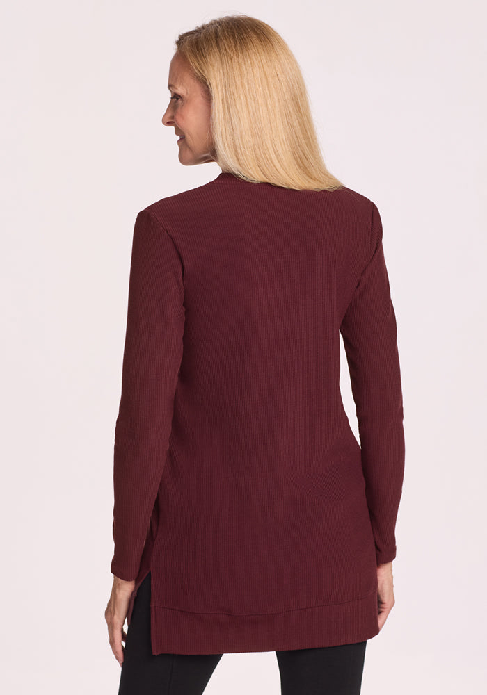 A woman with straight blonde hair wears the Woolx Evie Ribbed Tunic in Toasted Garnet and black pants, standing with her back to the camera against a plain light background.