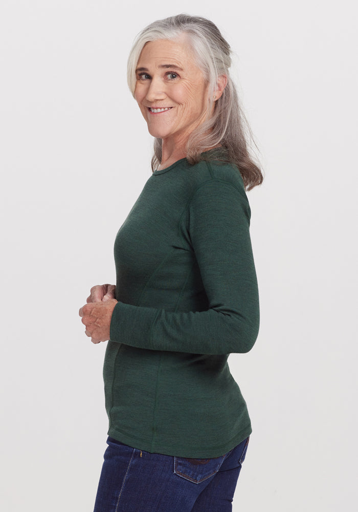 A smiling older woman with gray hair, in side profile, wears the Woolx Hannah Long Sleeve Top in Forest and blue jeans against a plain white background.